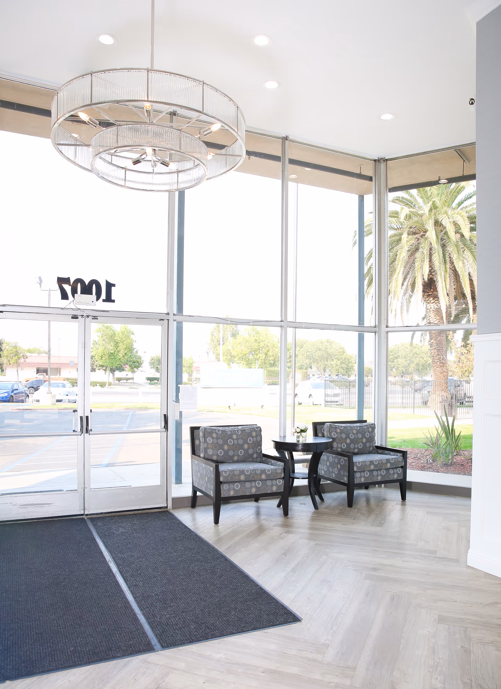 Bright lobby/entrance with large glass windows, two patterned armchairs and a small table beside double glass doors.