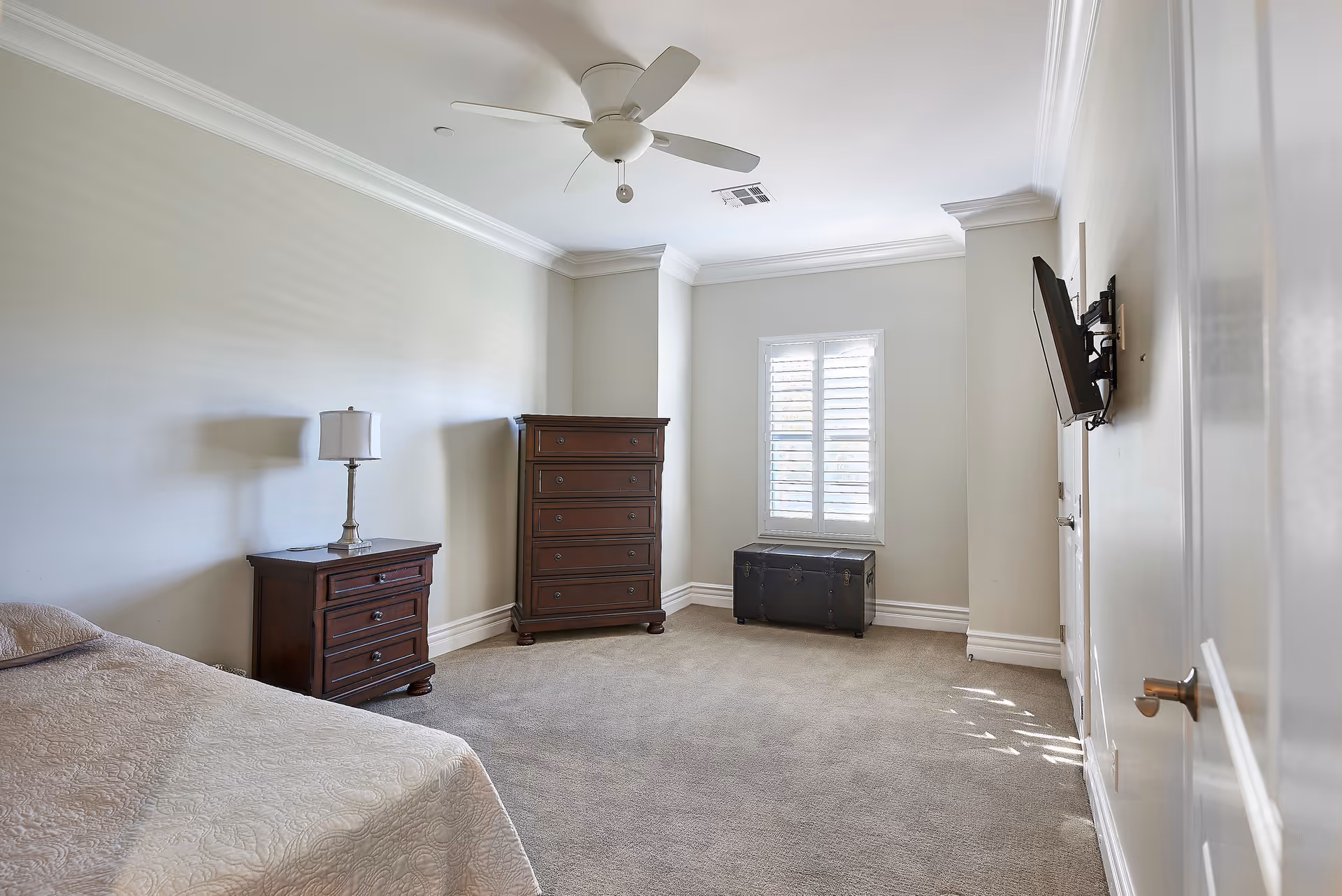 A bedroom with beige walls and carpeted floor featuring a bed with a beige quilt, a wooden nightstand with a lamp, a tall wooden dresser, a window with white shutters, a black trunk under the window, and a wall-mounted flat-screen TV.
