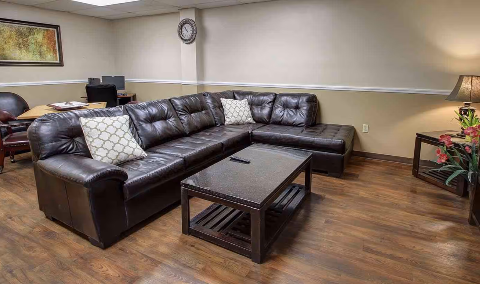 Cozy lounge room with a dark brown leather sectional sofa, central coffee table, side table with a lamp and flowers, and a desk area in the background.