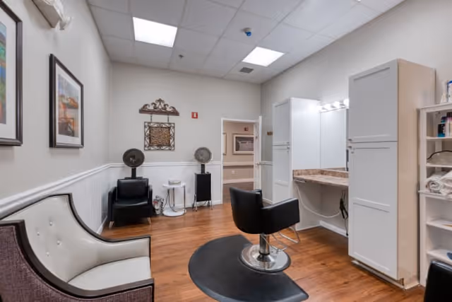 Interior view of a senior living facility's hair salon area featuring a styling chair in front of a large mirror with cabinets, a white cushioned armchair, two black chairs with hair dryers, framed artwork on the walls, and wood flooring.