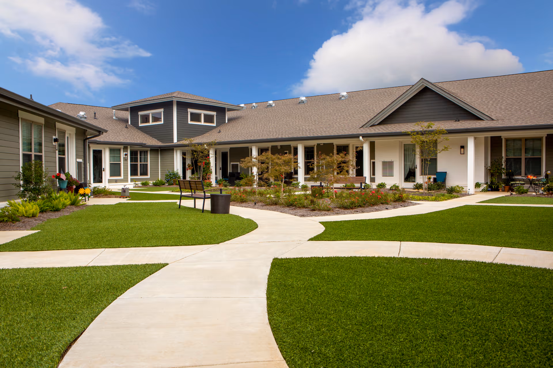 Outdoor courtyard area of a senior living facility with well-maintained green lawns, paved walkways, benches, and a garden with small trees and flowers. The building surrounding the courtyard has a modern design with large windows and a covered porch area under a partly cloudy blue sky.