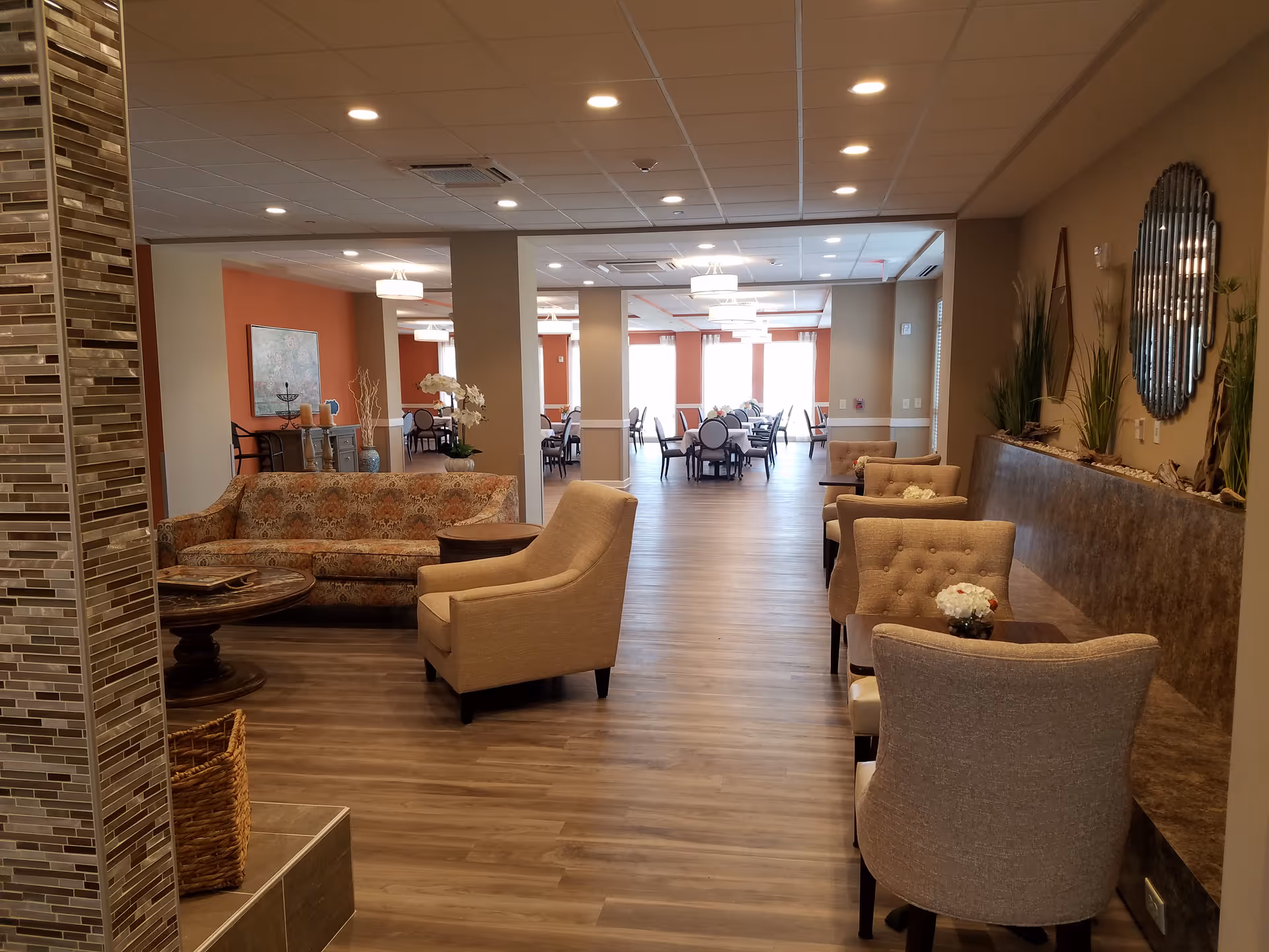 Spacious senior living lounge with sofas, armchairs, small tables, and a dining area visible in the background.