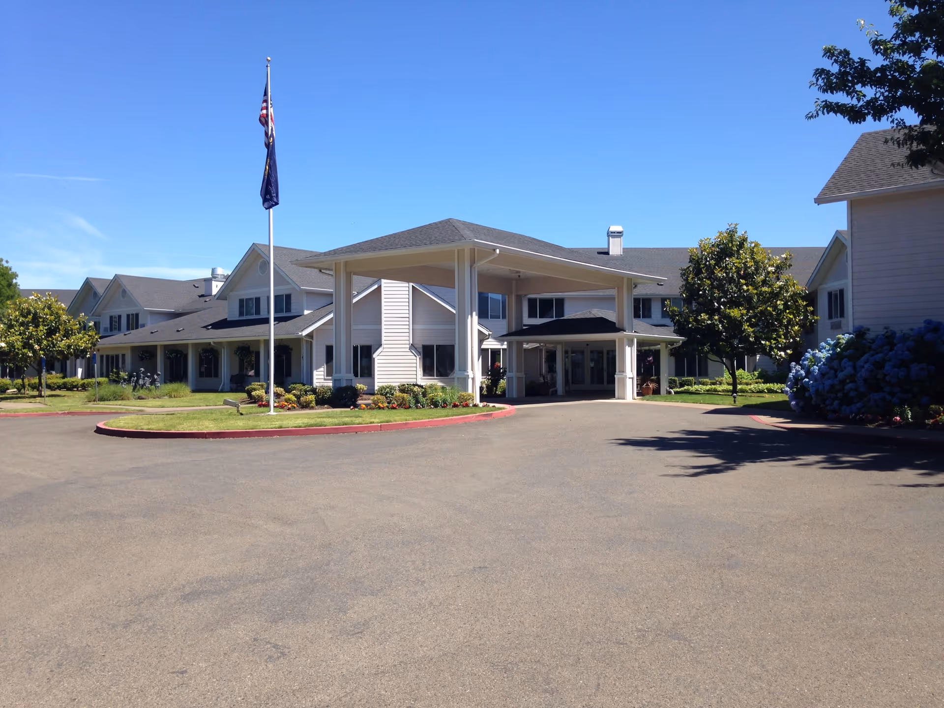 Exterior front view of Somerset Assisted Living facility showing a large covered entrance with a driveway, manicured landscaping including trees and bushes, and a flagpole with two flags against a clear blue sky.
