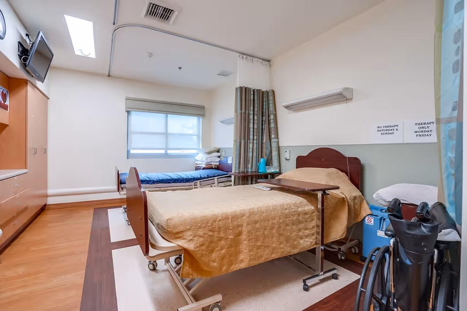 A senior care patient room with two beds, overbed tables, a wheelchair, and a curtained divider.