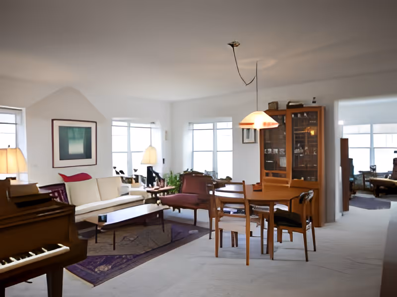A bright living room and dining area with white walls and carpet. The living room features a white sofa, wooden coffee table, purple armchairs, and a piano on the left. The dining area has a wooden table with four chairs and a hanging light fixture above it. A wooden cabinet with glass doors stands against the wall. Large windows allow natural light to fill the space.