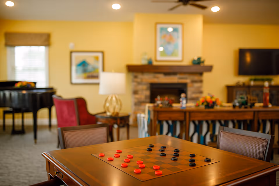 A cozy living room area in an assisted living facility featuring a wooden checkers table with red and black pieces set up for a game. In the background, there is a stone fireplace with a wooden mantle, a flat-screen TV mounted on the wall, a red armchair, a piano, and framed artwork on yellow walls. The room is warmly lit with ceiling lights and a table lamp.