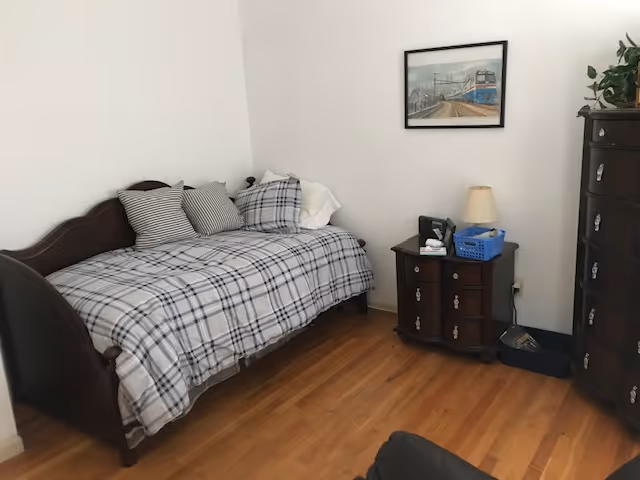 A small bedroom with a wooden daybed covered with a plaid blanket and three pillows. Next to the bed is a dark wooden nightstand with a lamp, a phone, and a blue basket. On the right side, there is a tall dark wooden dresser. A framed picture of a train hangs on the white wall above the nightstand. The floor is wooden.