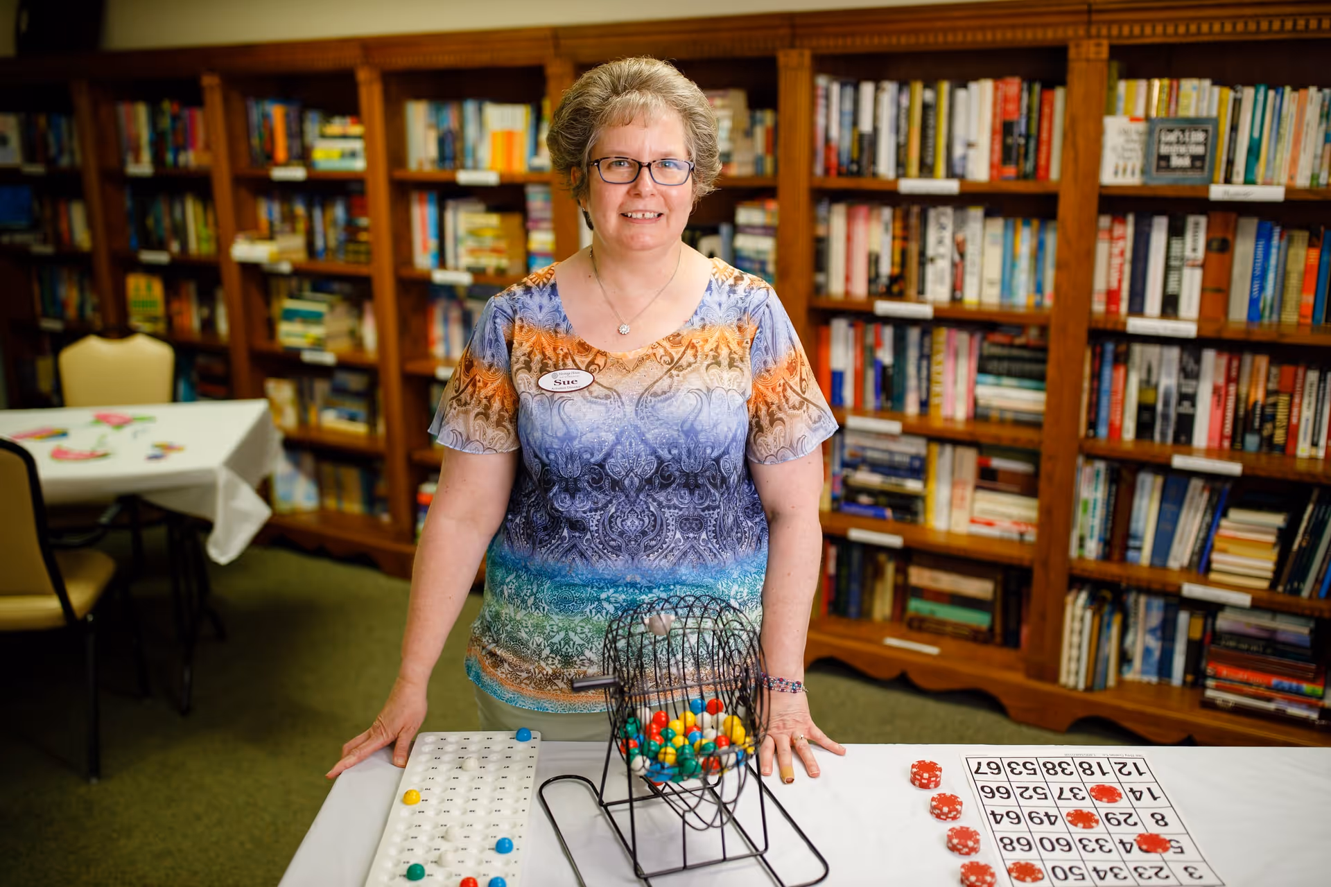 A woman wearing glasses and a colorful patterned shirt stands behind a table with bingo cards, chips, and a bingo cage in a room with wooden bookshelves filled with books.