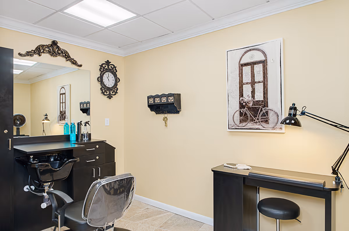 Interior view of a salon area in a senior living facility with a black salon chair and sink, a large mirror, hair care products on the counter, a decorative wall clock, a wall-mounted key holder, a desk with a black lamp, and a framed picture of a bicycle leaning against a door.