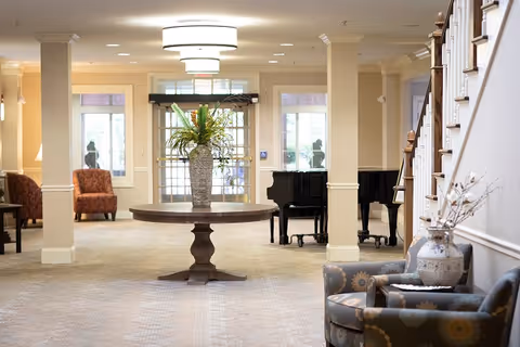 A spacious and well-lit common area with a round wooden table in the center holding a large vase with green and yellow flowers. The room features beige walls, carpeted floors, two upholstered chairs with floral patterns on the right, and a black grand piano near the back. There are large windows and glass doors letting in natural light, and a staircase is visible on the right side.