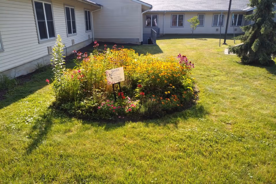 A sunny outdoor garden area at Riverwalk Village at The House of The Good Shepherd featuring a circular flower bed with various colorful flowers surrounded by green grass, adjacent to a beige building with multiple windows.