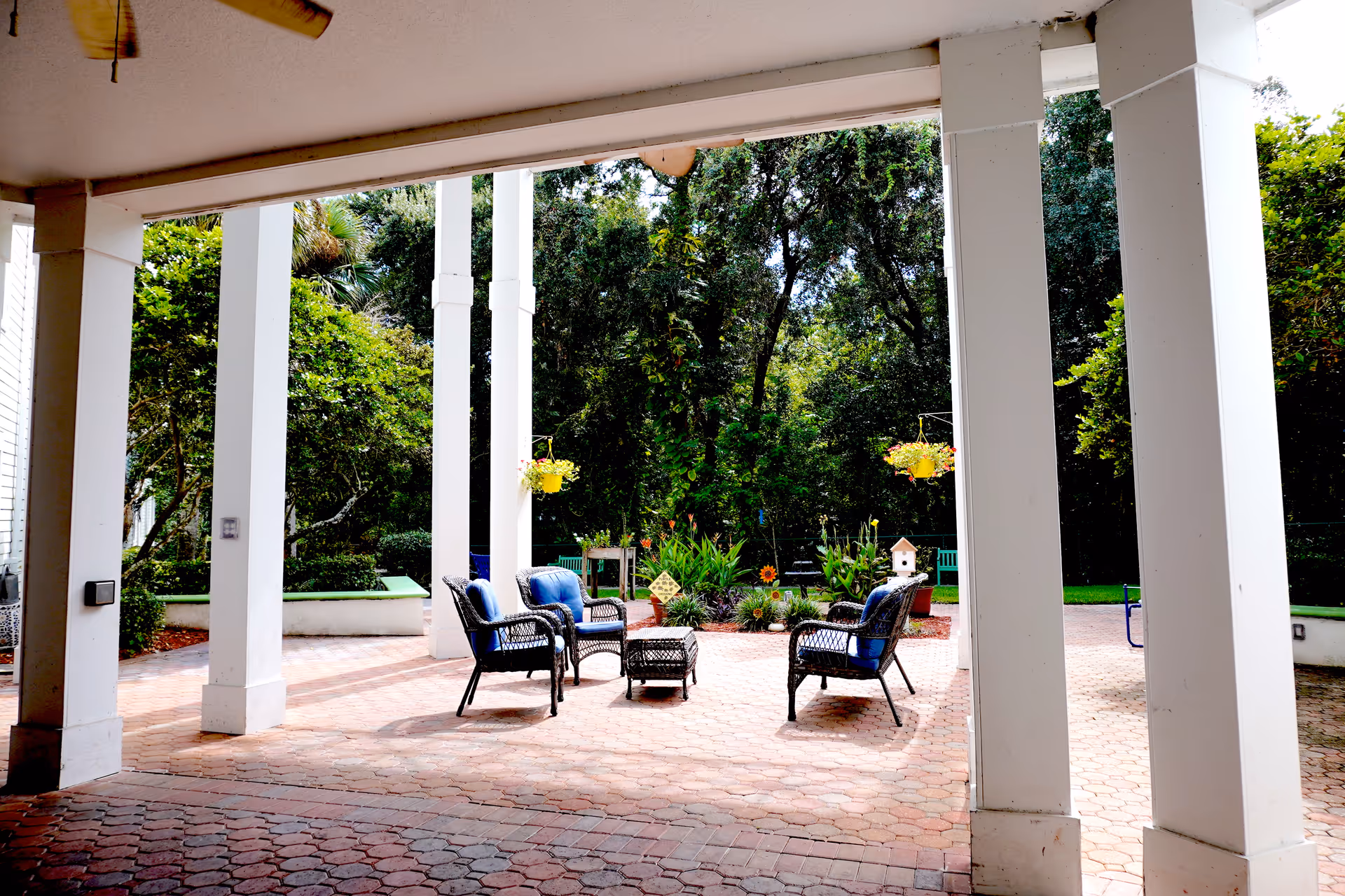 Outdoor patio area with four black wicker chairs with blue cushions arranged around a small black wicker table. The patio is paved with red bricks and surrounded by white columns. There are hanging yellow flower pots and a garden with various plants and trees in the background.