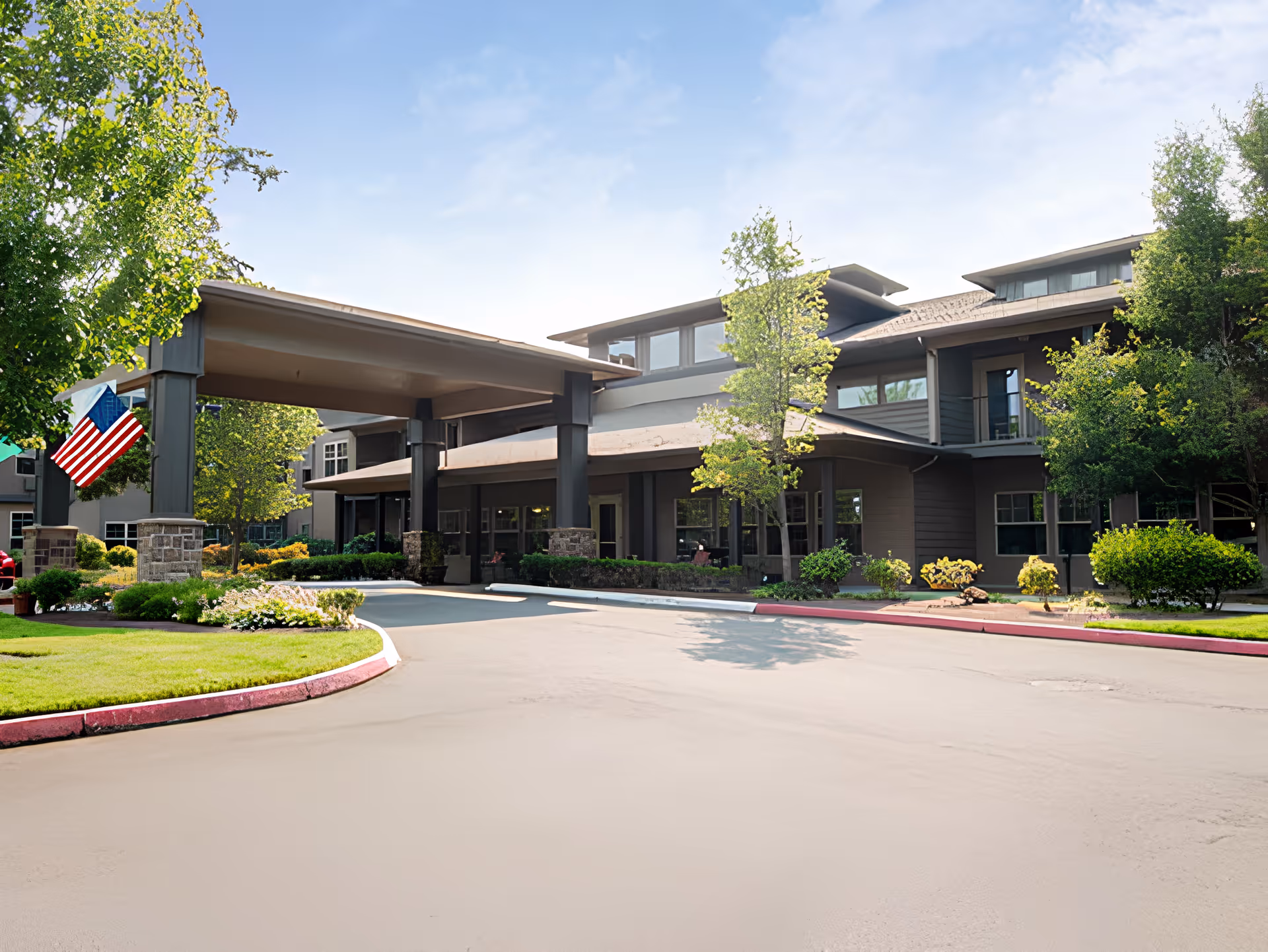 Exterior view of Port Townsend Senior Living by Cogir showing the entrance with a covered driveway, surrounded by well-maintained landscaping including green trees and bushes under a partly cloudy sky.