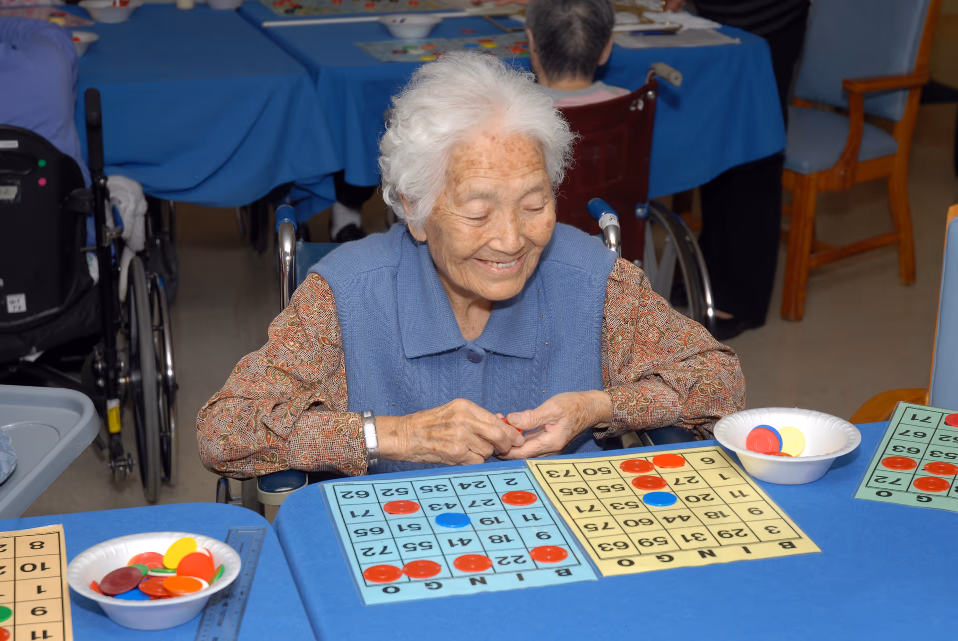 An elderly woman with white hair sitting in a wheelchair at a table covered with a blue tablecloth, smiling while playing bingo with colorful chips on bingo cards. Other elderly individuals are also seated at tables in the background.