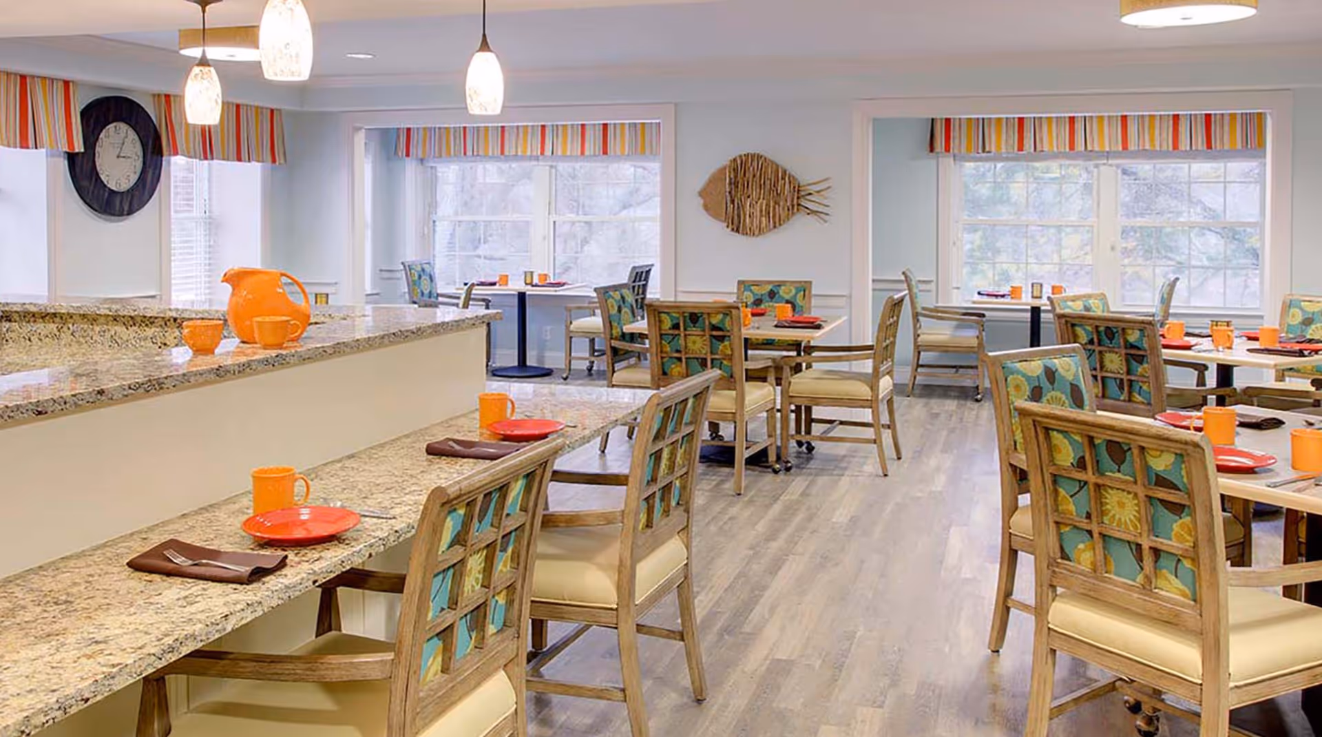 A bright dining room with multiple wooden tables and chairs featuring colorful patterned cushions. The tables are set with orange mugs, red plates, and brown napkins. There is a granite countertop with additional seating in the foreground, and large windows with striped valances letting in natural light. A decorative fish wall art is mounted on the far wall.