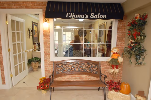 Interior view of a salon area named 'Eliana's Salon' with a decorative wooden bench in front of a brick wall. The salon window shows a person inside, and the area is decorated with autumn-themed items including flowers, a scarecrow doll, and a pumpkin. There is a door with glass panels to the left and a wall-mounted light fixture above the bench.