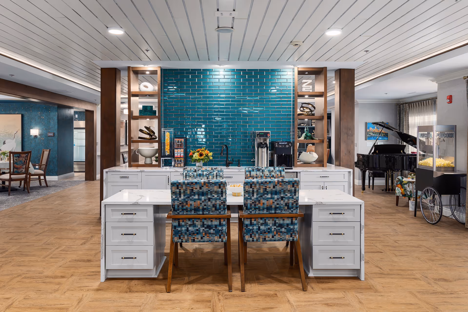 A modern common area with a white island table surrounded by four patterned chairs. Behind the table is a teal tiled wall with a coffee station, a sink, and decorative shelves. To the right, there is a popcorn machine and a black grand piano. To the left, there is a dining area with wooden chairs and a large painting on the wall. The floor is wood-patterned, and the ceiling has white panels with recessed lighting.