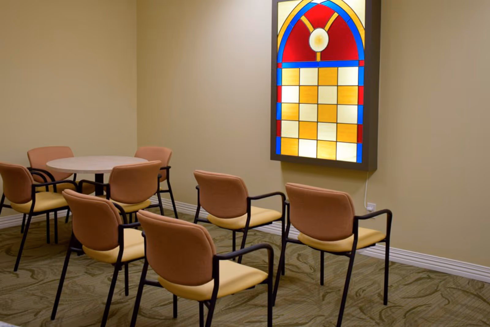 A small room with six chairs arranged in two rows facing a wall-mounted stained glass window with a colorful geometric design. There is also a round table with three chairs around it in the corner. The walls are beige and the floor has a patterned carpet.