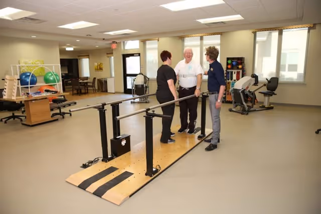 Three people standing and talking in a spacious rehabilitation or physical therapy room with parallel bars for walking exercises, exercise balls, and various fitness equipment in the background.