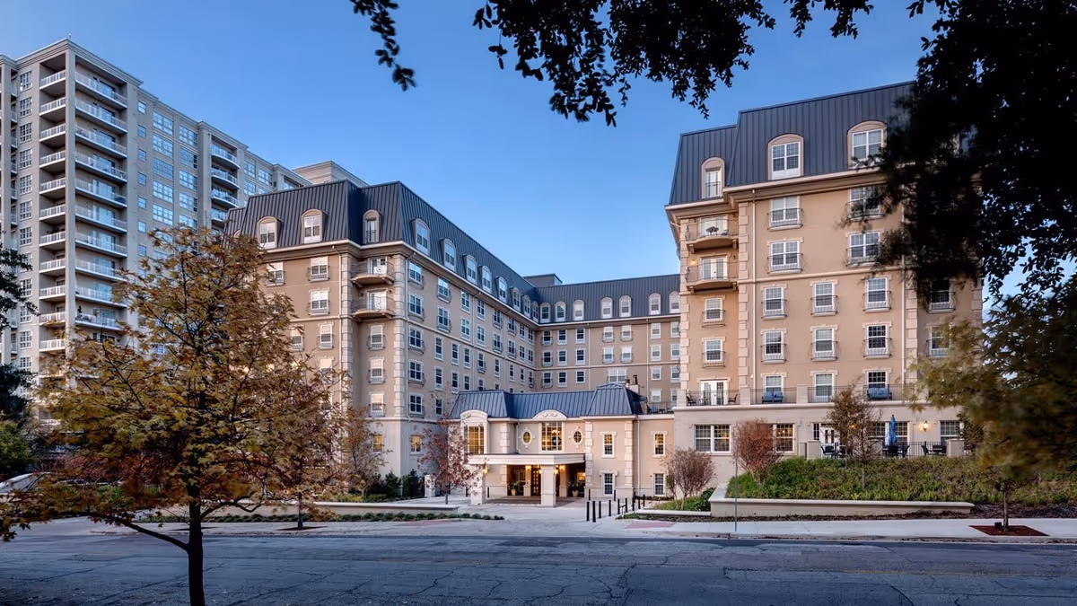 Exterior view of Belmont Village Senior Living Turtle Creek, a multi-story residential building with beige walls and a dark roof, surrounded by trees and landscaping under a clear blue sky.