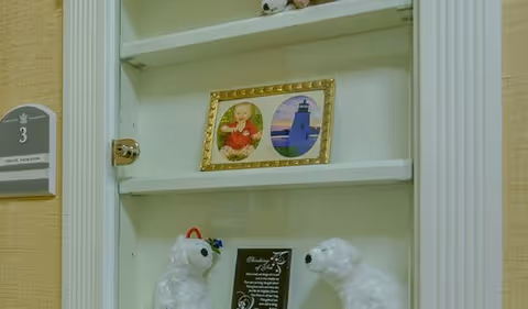 A white recessed wall shelf displaying decorative items including a framed photo of a baby and a lighthouse, two white polar bear figurines, and a small black plaque with text. To the left of the shelf is a room number sign labeled '3'.