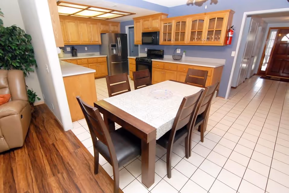 Interior view of a kitchen and dining area in a senior living facility. The kitchen features wooden cabinets, a stainless steel refrigerator, a black stove, and a microwave. The dining area has a wooden table with six chairs and a white table runner. The floor is tiled, and there is a hallway leading to a wooden front door. A beige recliner and a green plant are partially visible on the left side.