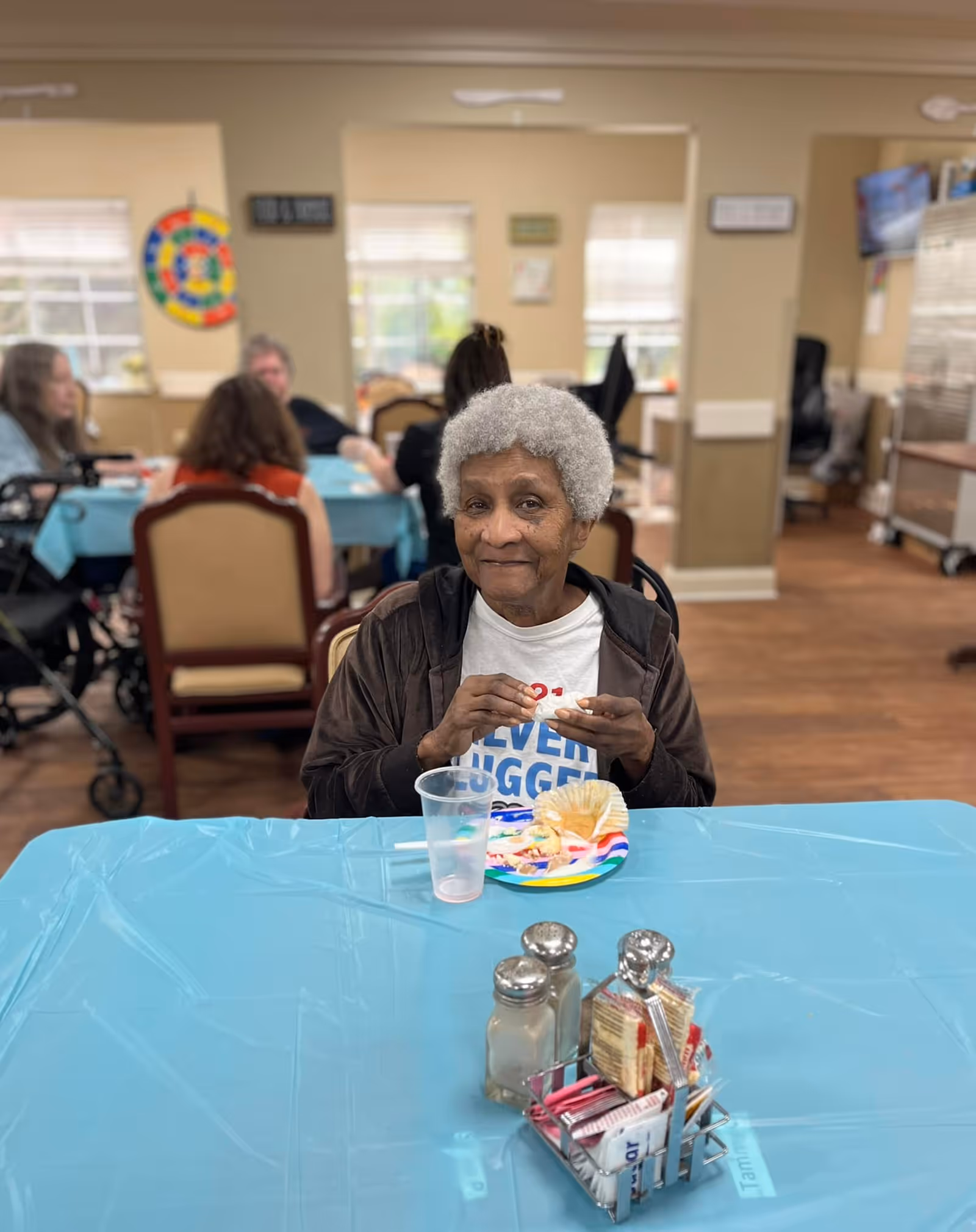 An elderly woman with gray hair sitting at a table covered with a blue tablecloth in a dining area. She is holding a piece of food and smiling slightly. On the table in front of her is a colorful plate with food, a clear plastic cup, and a small container holding salt, pepper, and packets of condiments. In the background, other elderly people are seated at tables, and the room has beige walls and windows letting in natural light.