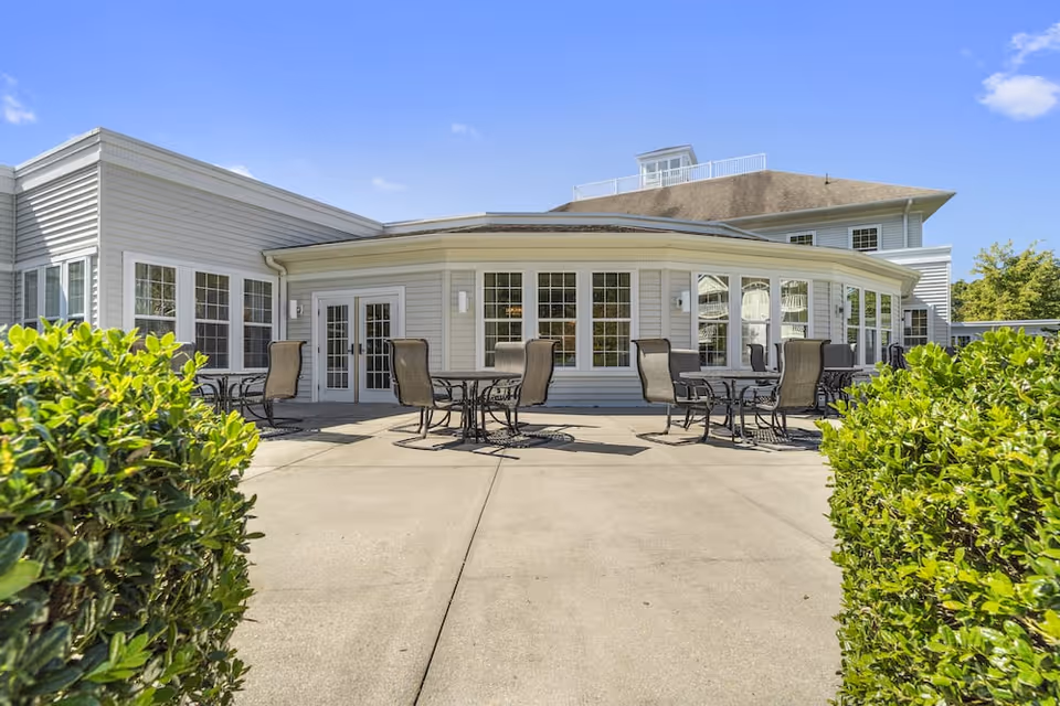 Outdoor patio area at Lakeside At Mallard Landing with several round tables and chairs arranged on a concrete surface, surrounded by green bushes and a building with large windows and white siding in the background under a clear blue sky.