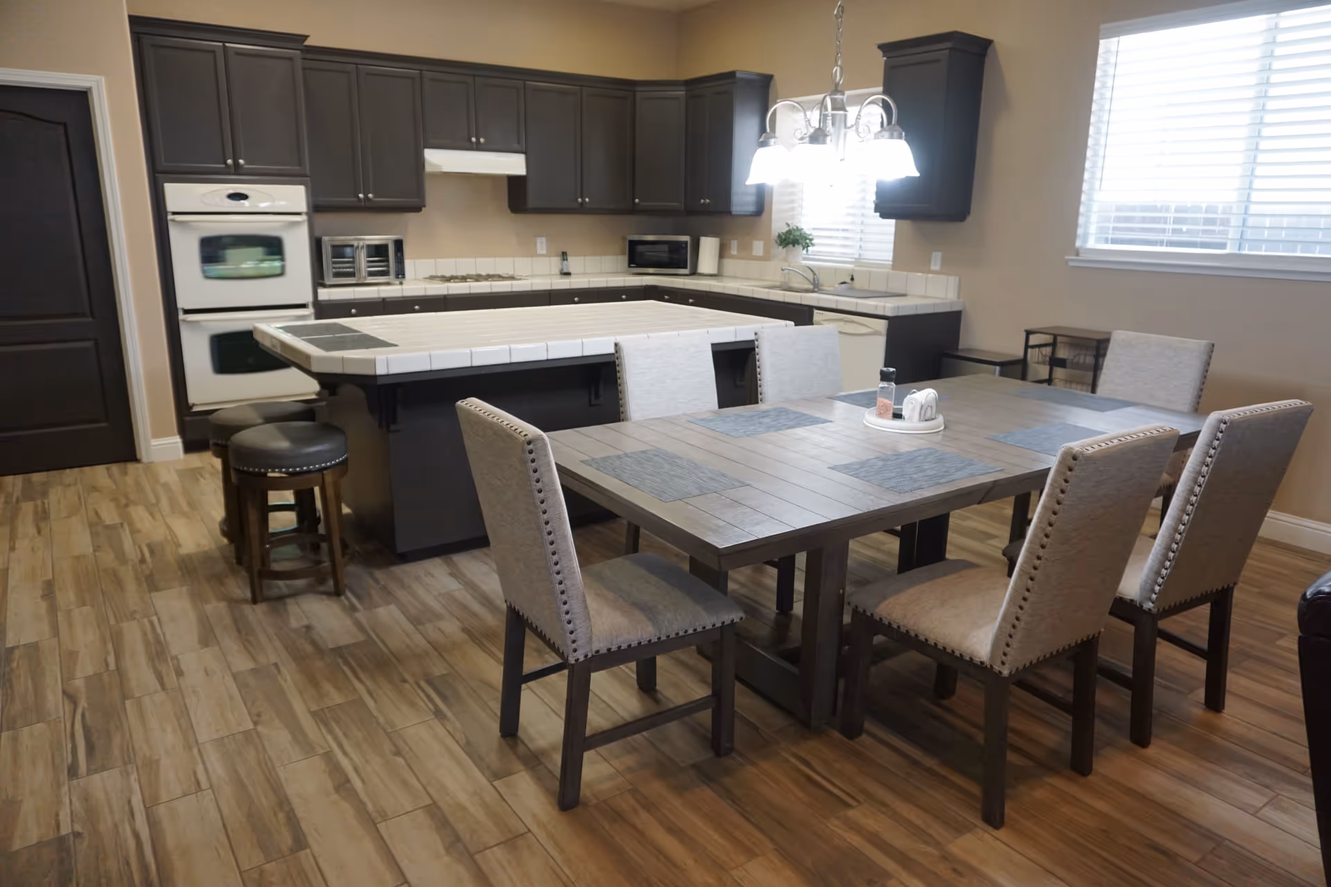 A modern kitchen and dining area with dark wood cabinets, a white tiled countertop island with three stools, a wooden dining table with six upholstered chairs, placemats, and a condiment set. The room has wood-look flooring and a window with blinds letting in natural light.
