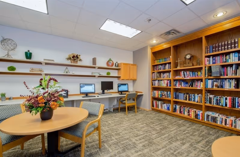 A cozy room with a round wooden table and four chairs, a large wooden bookshelf filled with books, and a desk area with two computer monitors. The walls have decorative shelves with various ornaments and plants. The room is well-lit with ceiling lights and has a carpeted floor.