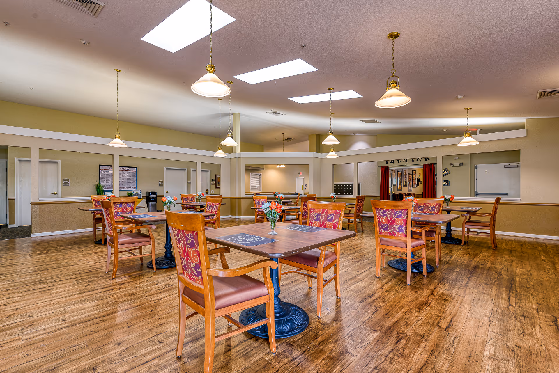 Spacious dining/activity room with multiple wooden tables and patterned chairs under pendant lights and skylights.