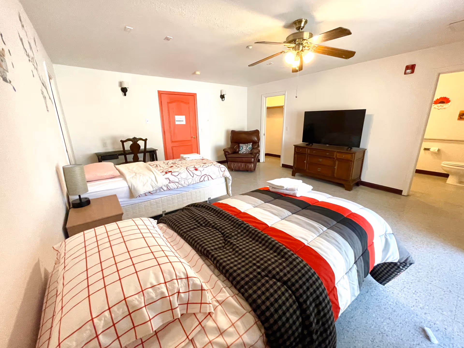 Bright assisted-living bedroom with two single beds, a dresser-mounted TV, recliner, ceiling fan, and an open bathroom door.