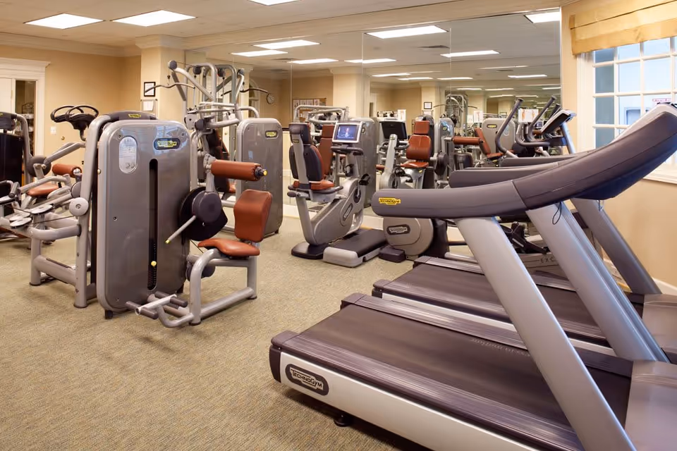 A well-lit fitness room with various exercise equipment including treadmills, stationary bikes, and strength training machines. The room has beige walls, a large mirror along one wall, and a window with a yellow valance.