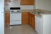 A clean kitchen with wooden cabinets, a white electric stove with oven, a white refrigerator, a white dishwasher, and a double sink with a countertop. The floor is light-colored and the walls are plain.
