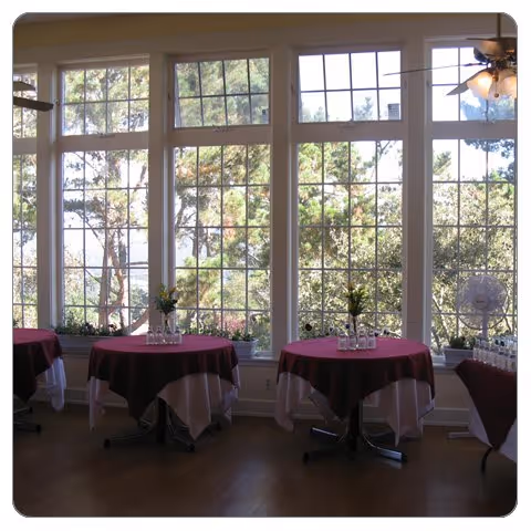 Sunlit room with large paned windows and small round tables covered with burgundy and white tablecloths.