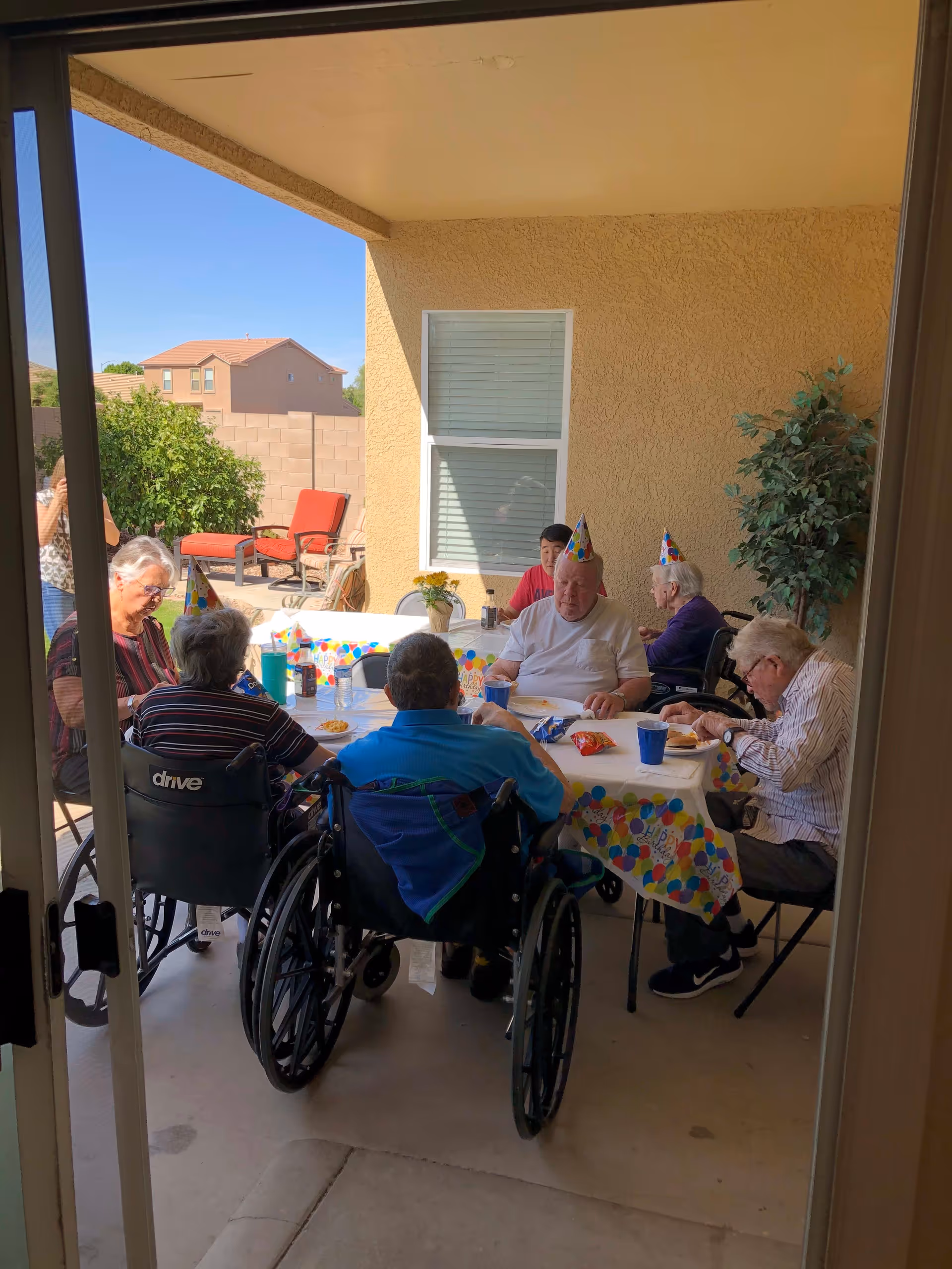 A group of elderly people, some in wheelchairs, sitting around a table outdoors under a covered patio. They are wearing colorful party hats and appear to be celebrating a birthday with snacks and drinks on the table. There is a potted plant and patio furniture in the background, with a clear blue sky and neighboring houses visible.
