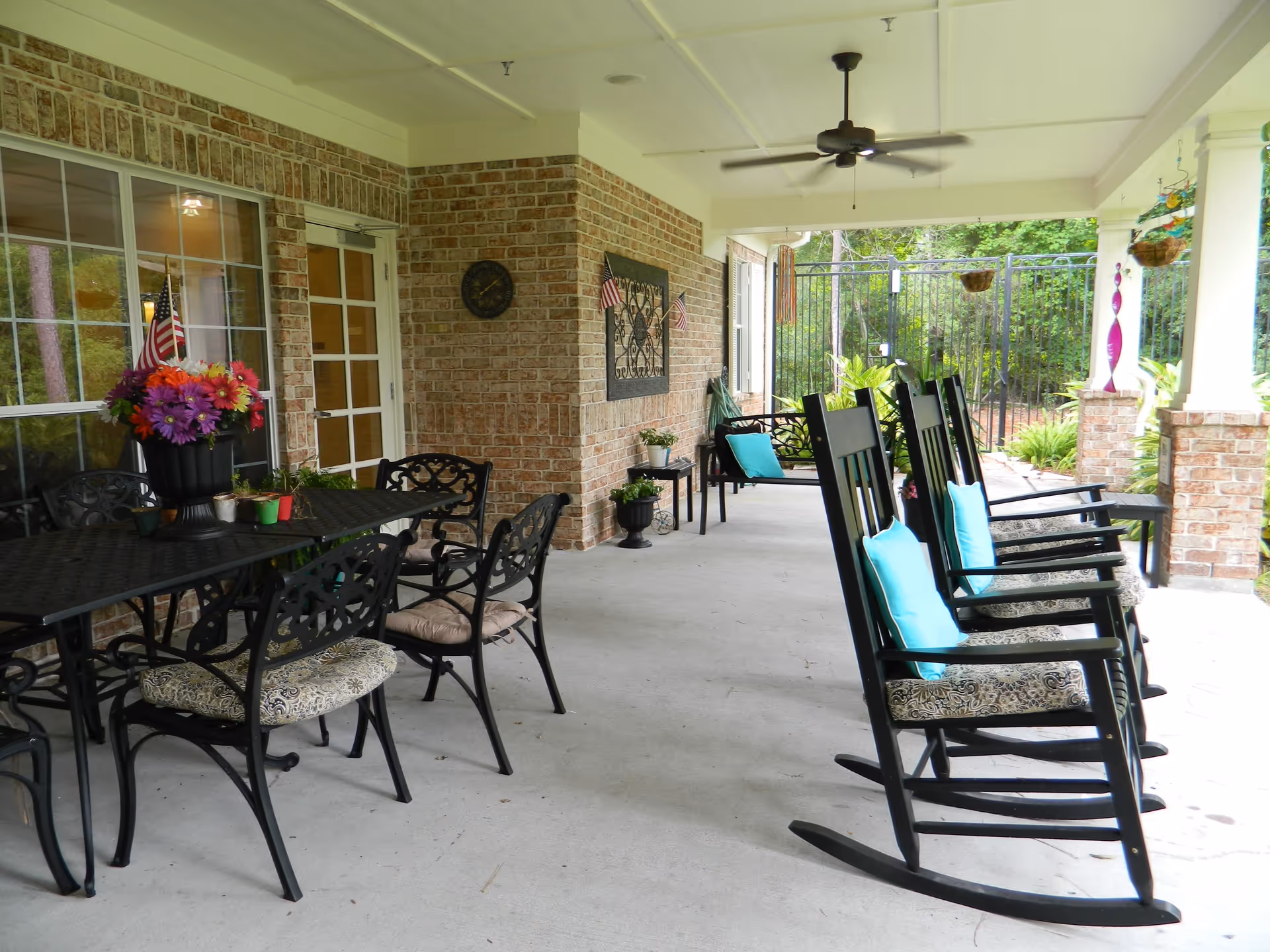 Covered outdoor patio area with black metal dining table and chairs, a vase of colorful flowers on the table, several black rocking chairs with blue cushions, brick walls, ceiling fan, and greenery visible beyond a black metal fence.