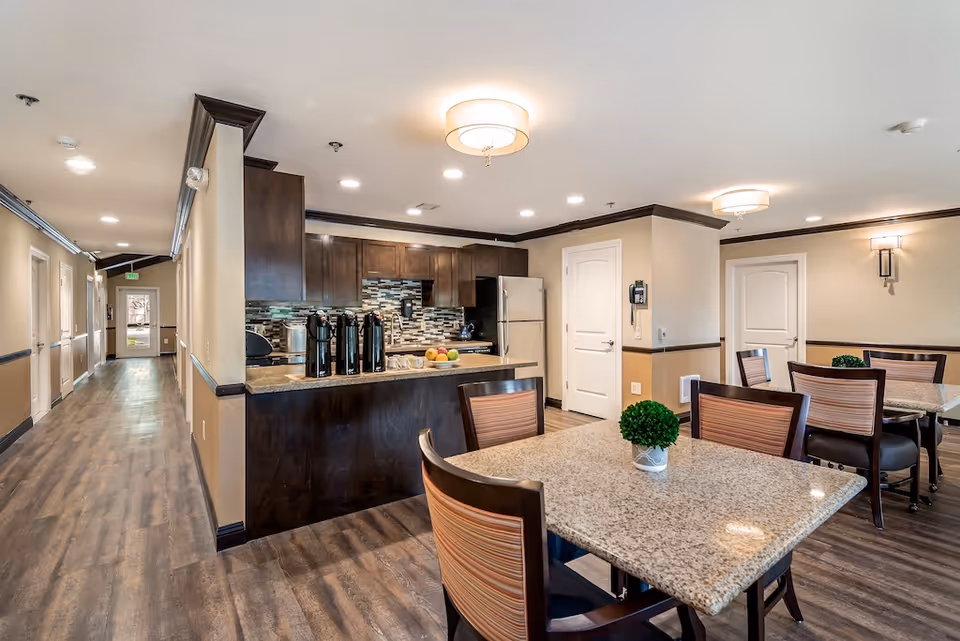 Interior view of a senior living facility dining area and kitchen at Pacifica Dogwood Cottage. The image shows a granite dining table with chairs and a small green plant centerpiece. In the background, there is a kitchen with dark wood cabinets, a stainless steel refrigerator, and a countertop with coffee dispensers and fruit. The hallway extends to the left with doors along the wall and an exit door at the end.