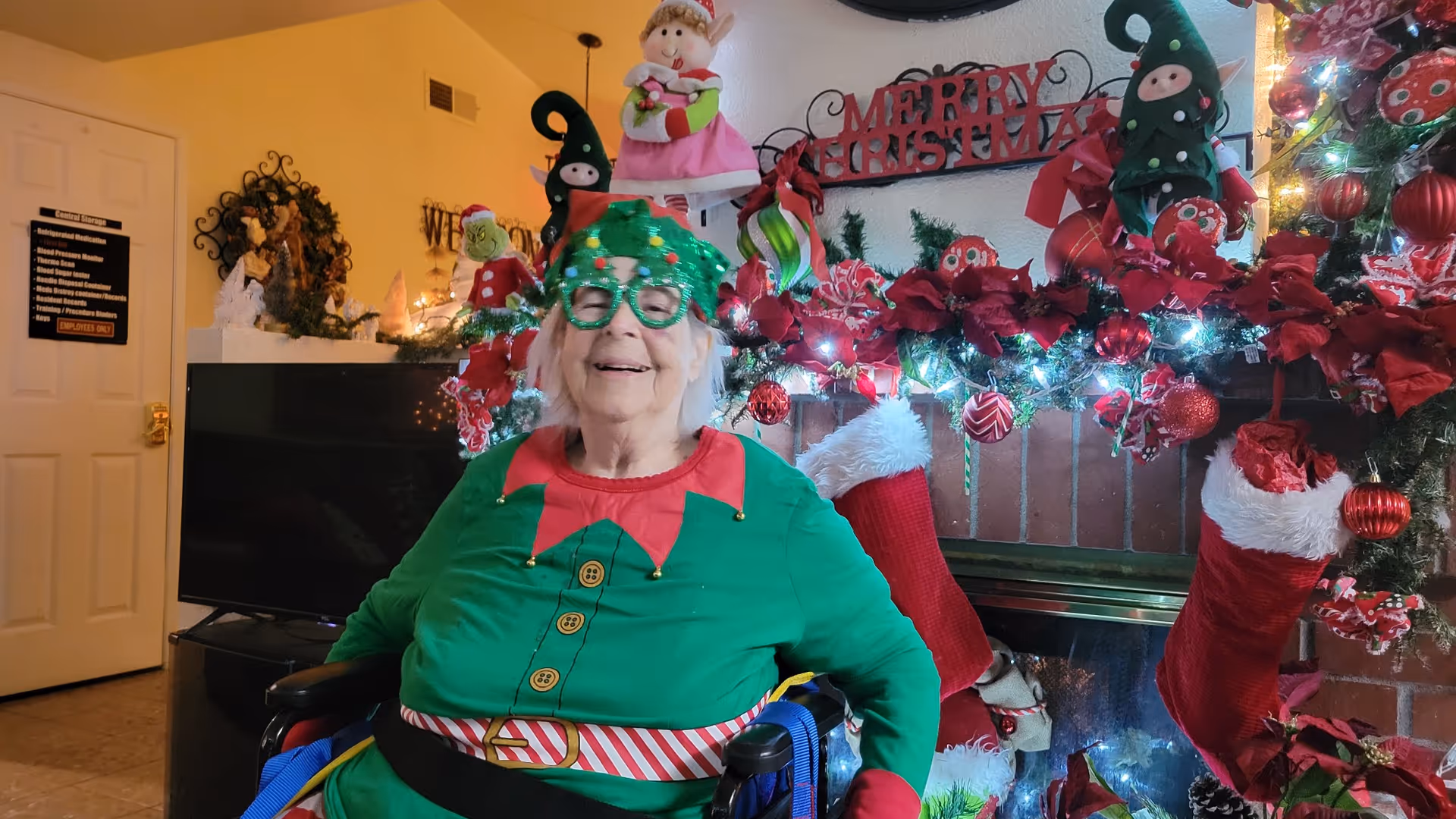 An elderly woman dressed in a green elf costume with festive glasses sits in a wheelchair in front of a decorated fireplace. The fireplace mantel is adorned with Christmas decorations including stockings, garlands with lights, poinsettias, and holiday figurines. A 'Merry Christmas' sign is displayed above the mantel. The room has warm lighting and additional holiday decorations are visible in the background.