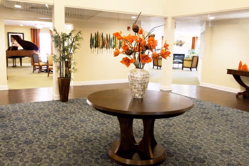 Interior view of a senior living facility lobby area with a round wooden table in the center holding a vase with orange flowers. The room has patterned carpet, light-colored walls, and decorative wall art. In the background, there is a seating area with chairs and a piano visible.