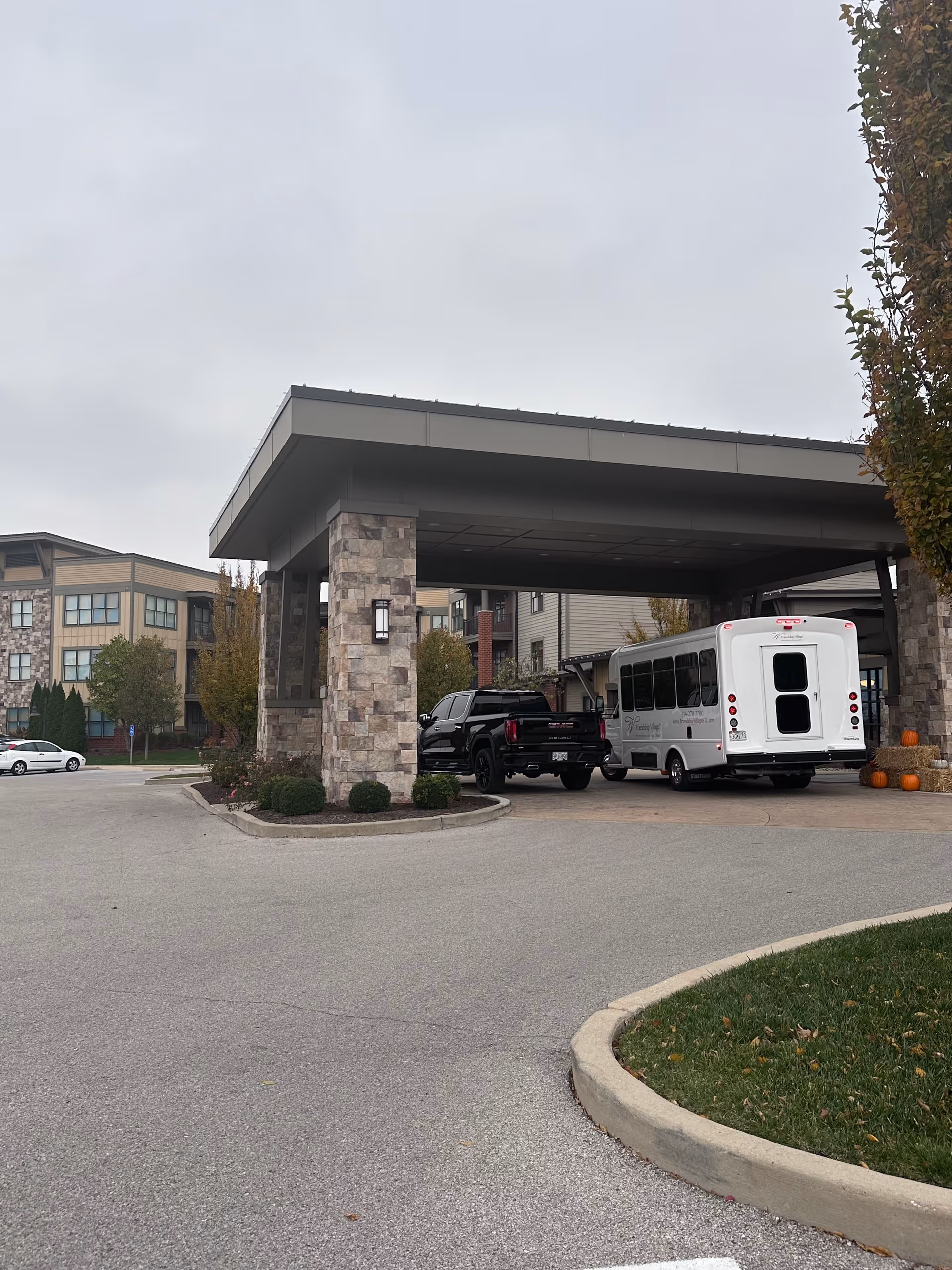 Entrance area of Friendship Village Sunset Hills showing a covered drop-off zone with stone pillars. A black pickup truck and a white shuttle bus with the facility's name are parked under the canopy. The building exterior is visible in the background with trees and a cloudy sky.