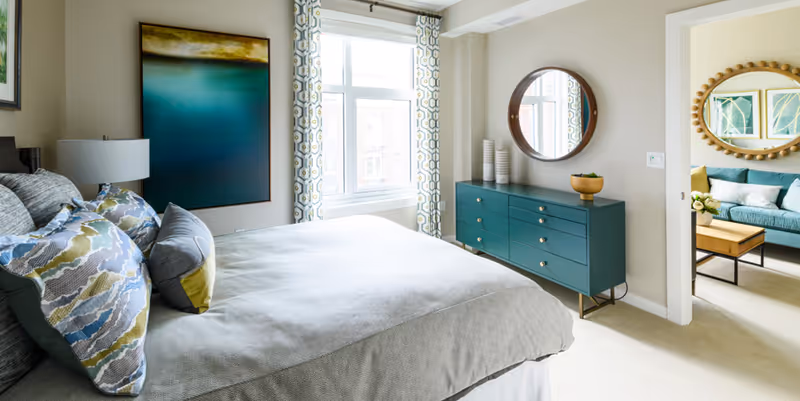 Bright bedroom with a bed, patterned pillows, a teal dresser with round mirror, window, and a view into an adjacent living area.
