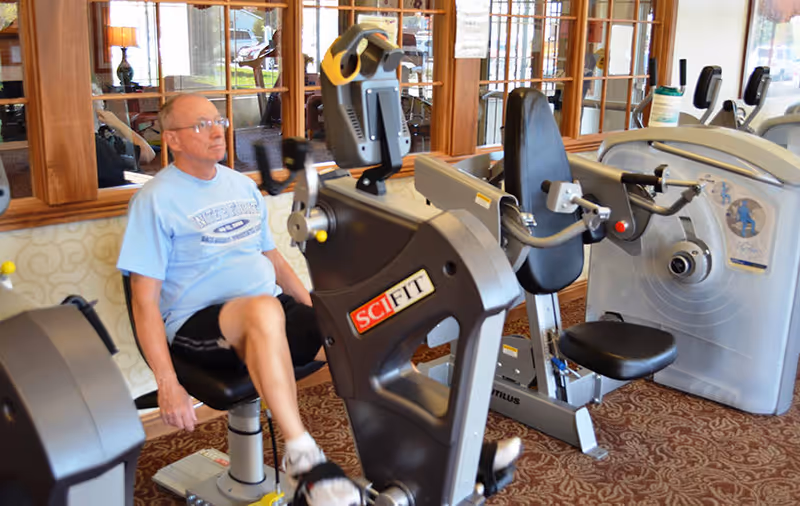 An elderly man wearing a light blue t-shirt and black shorts is exercising on a SciFit stationary leg exercise machine in a fitness room with carpeted floor and large windows with wooden frames.