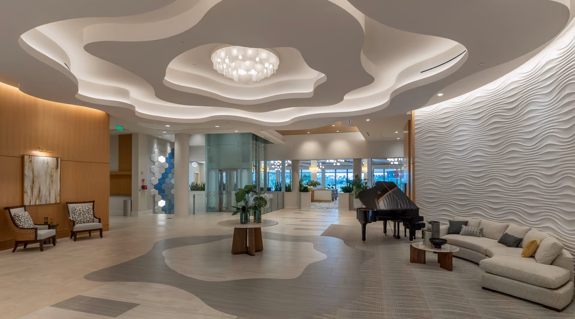 A spacious and modern interior lobby area with a wavy ceiling design and a large chandelier. The room features a curved beige sofa with cushions, a black grand piano, a round wooden table with plants, and two armchairs against a wooden wall with artwork. The far end shows glass doors and windows with natural light coming through.