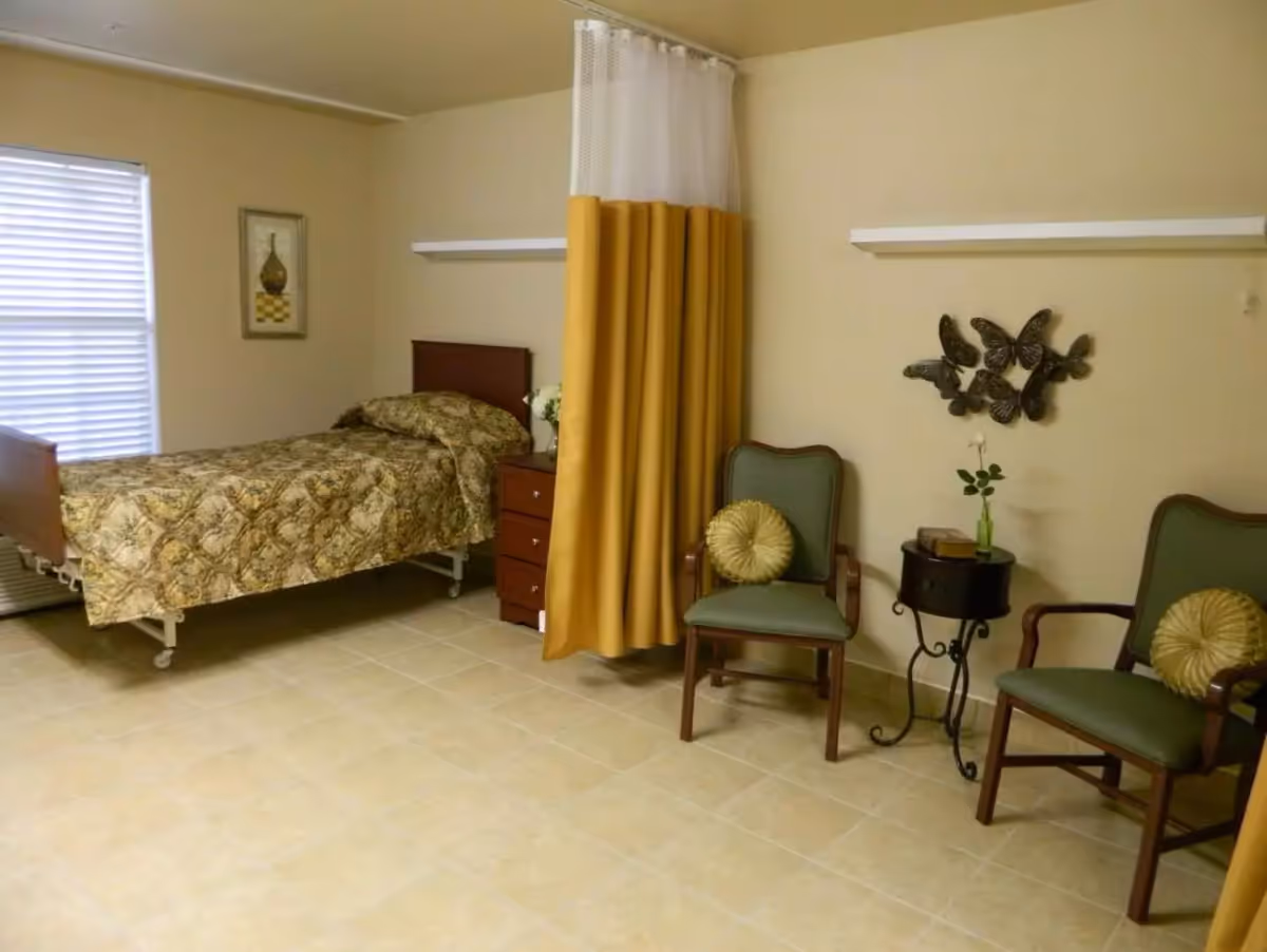 A nursing facility bedroom with a single adjustable bed, a yellow privacy curtain, bedside drawers, two armchairs and a small side table with wall decor.