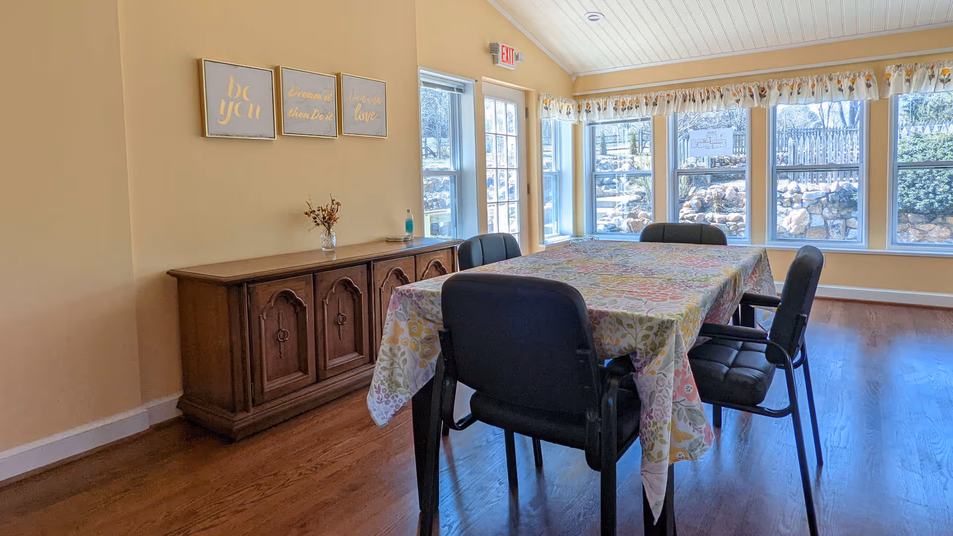 A bright dining room with a rectangular table covered by a floral tablecloth surrounded by four black cushioned chairs. There is a wooden sideboard against the wall with a small vase of dried flowers and a bottle of hand sanitizer on top. Three framed inspirational quotes hang above the sideboard. Large windows and a glass door let in natural light and provide a view of a stone wall and garden outside.