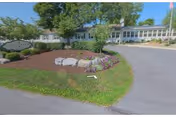 Exterior view of a single-story nursing center building with a landscaped garden featuring rocks, mulch, and flowering plants in front. Trees and shrubs surround the building, and a paved driveway curves in front of the entrance under a clear blue sky.