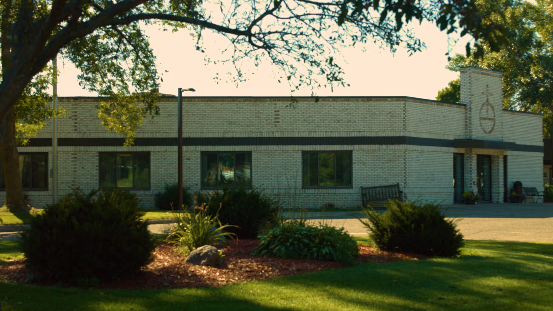 Front exterior of a single-story brick rehabilitation center with landscaping, benches, and a tree-lined lawn.