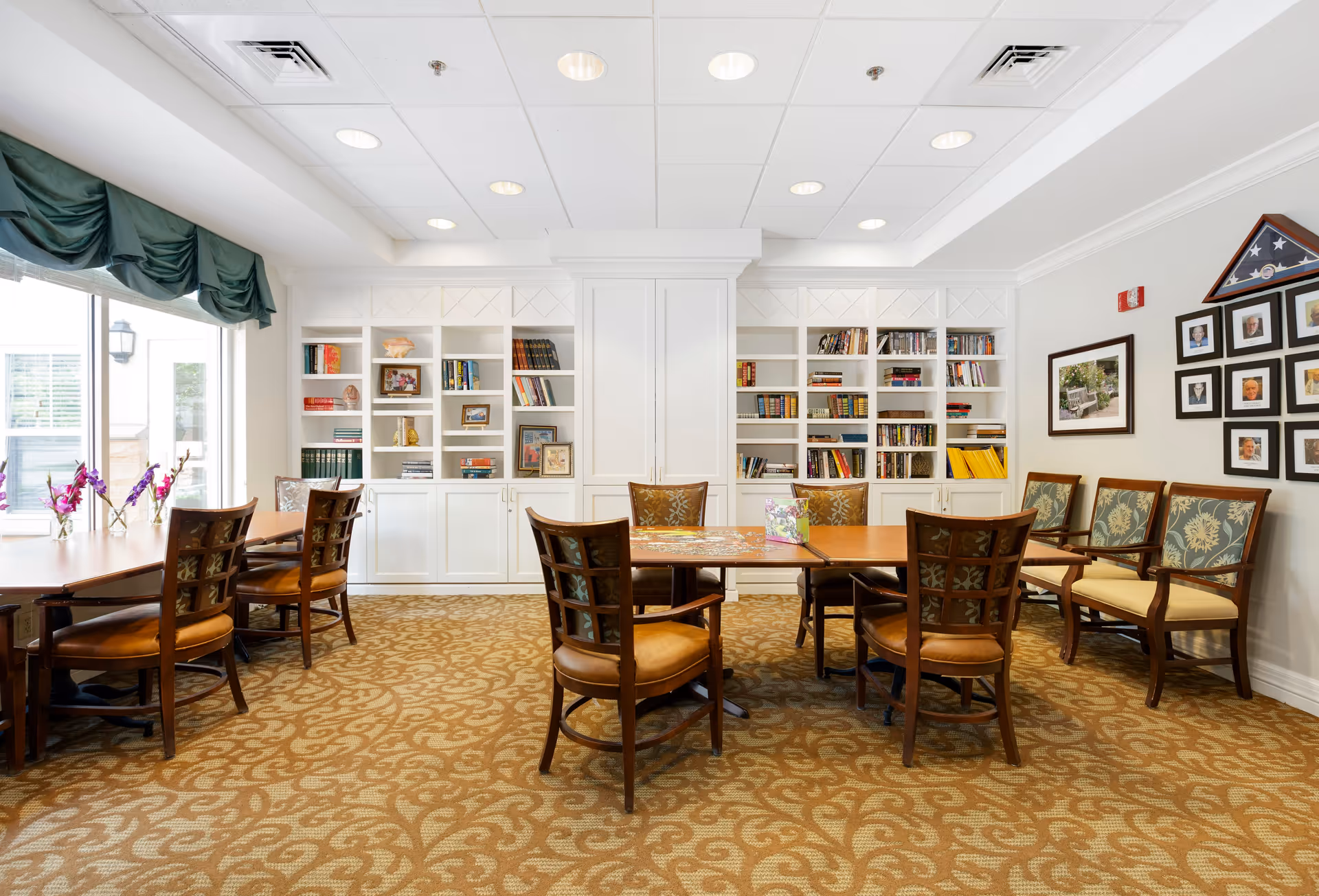 A well-lit room with patterned carpet, wooden tables and chairs arranged for group seating. The back wall features white built-in bookshelves filled with books and decorative items. On the right wall, there are framed photographs and a folded American flag displayed in a triangular case. Large windows with green valances allow natural light into the room.