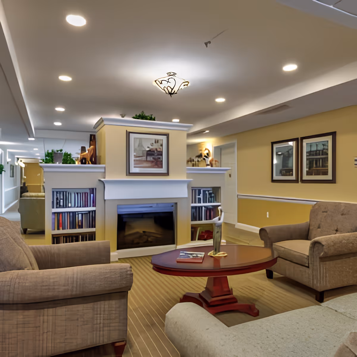 A cozy living room area with beige upholstered armchairs and a sofa arranged around a wooden coffee table. The room features a fireplace with built-in bookshelves on either side, decorated with plants and artwork above the mantel. The walls are painted yellow with white trim, and framed pictures hang on the wall. The ceiling has recessed lighting and a decorative light fixture.